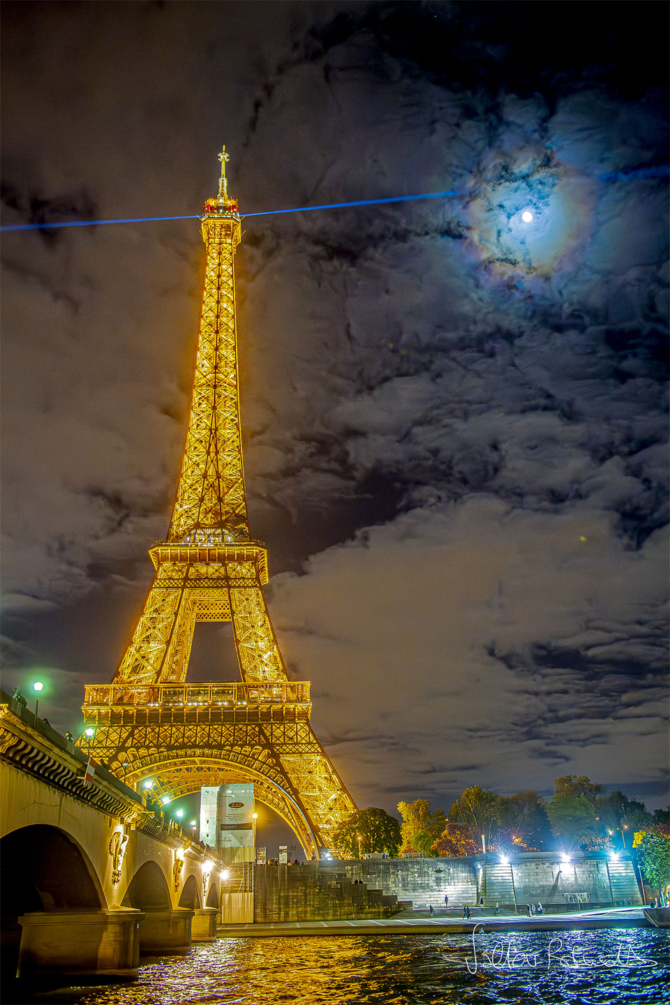 Po lewej stronie zdjęcia widoczna jest słynna, paryska wieża Eiffela, podświetlona na złoty kolor. Z jej szczytu wydobywa się promień niebieskiego lasera.
Na niebie widać pojedyncze chmury. Przez chmury widoczny jest Księżyc, otoczony kolorowymi pierścieniami: koroną słoneczną.
Więcej szczegółowych informacji w opisie poniżej.